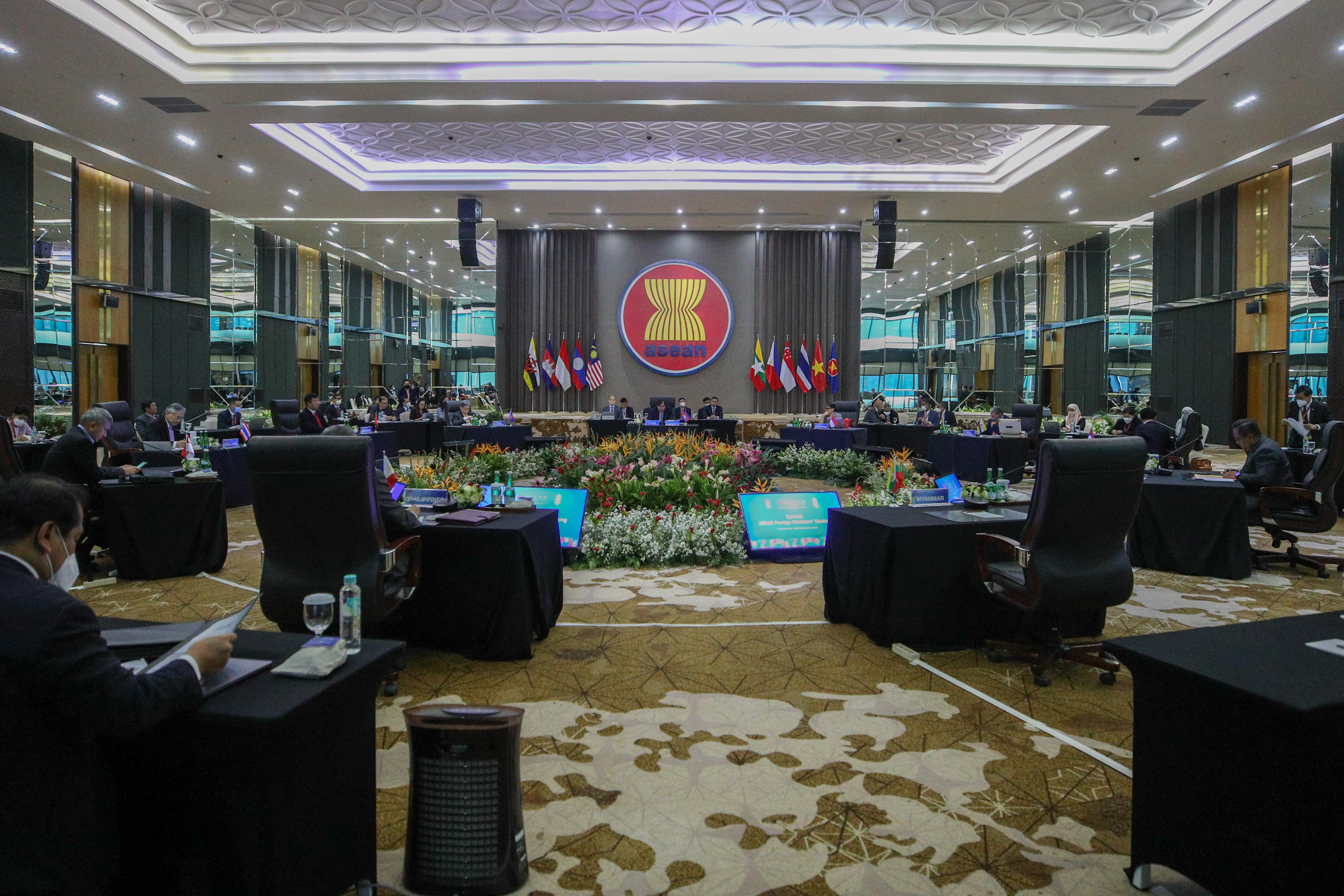 ASEAN meeting room with flag backdrop, floral displays, delegates seated at tables, and patterned carpeting.